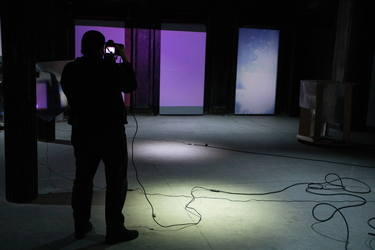Image of a dimly-lit basement, a figure holding a video camera with a rig on their shoulder takes footage of the back wall, which has blue and purple light projected onto it.