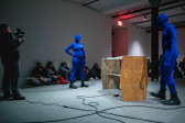 Three figures perform in front of an audience. Two figures in blue morph suits face off with a wooden table between them, while a third figure takes video footage of the scene.