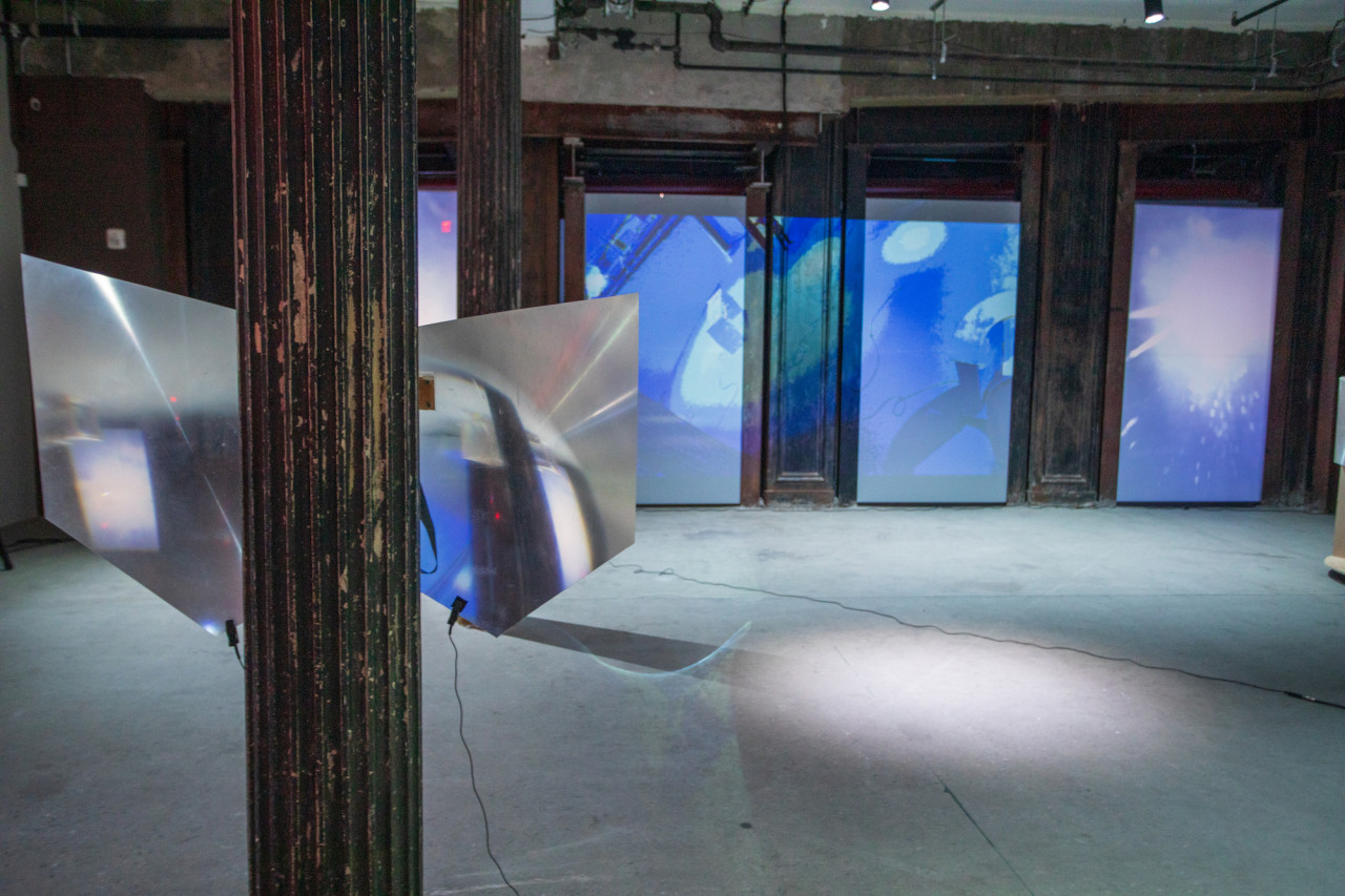 Image of a basement with a sculpture made of two fresnel lenses cut in the shape of wings hung on the back of a pillar to the left of the image. Along the back wall a blue projected image of live CCTV footage of the basement is displayed.