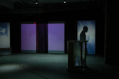 Image of a dimly lit basement. Along the back wall, on the right, a shadow of a figure in profile is cast onto a blue and purple projected screen.