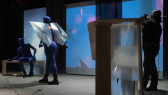 Two figures in blue morph suits face off, with one figure sitting down on a wooden box looking up at the other figure who stands above them wearing wings made of fresnel lenses. All the way on the right a figure takes video footage of the scene.