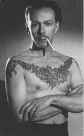 A slightly grainy image of a man standing shirtless with his arms crossed. He has a large, floral tattoo spanning from shoulder to shoulder across his chest, and a cigarette in his mouth.