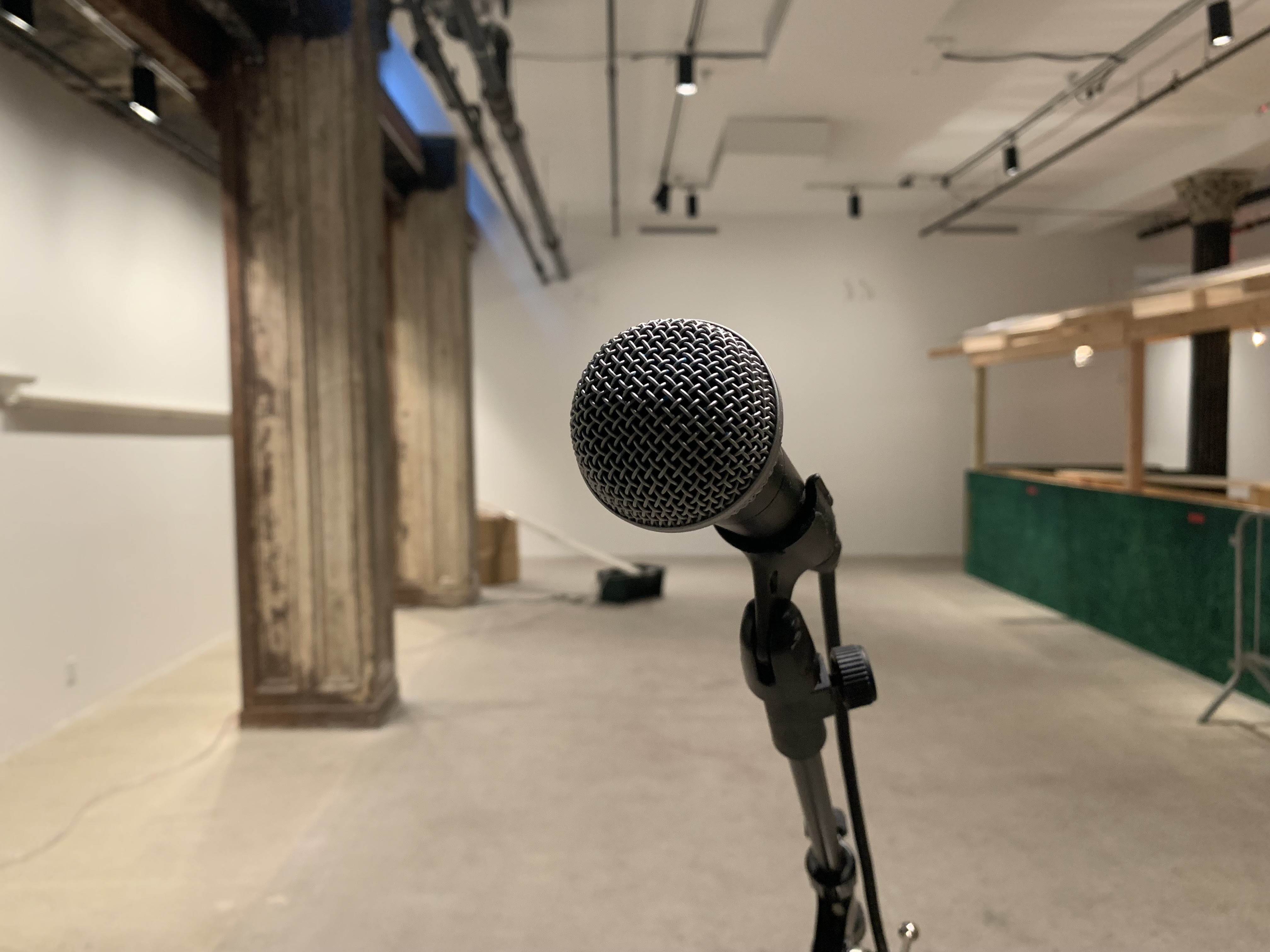 A crisp image of a microphone centered in front of an out of focus background. Behind the microphone are steel columns to the left, and a green wooden structure to the right.