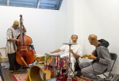 Photograph of three musicians in a room with a red carpet. The figure on the left is playing an upright bass, the center figure is seated behind a drum set, and the figure on the left holds a guitar with their back turned to the camera.