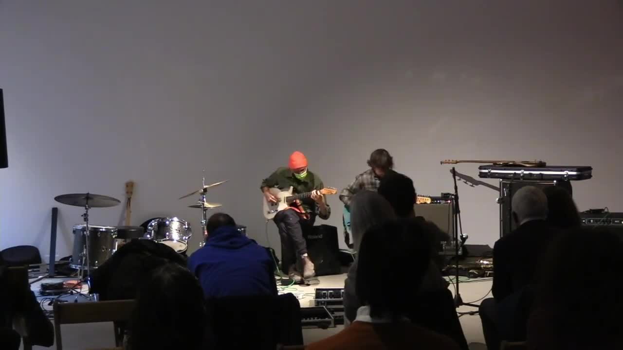 A color video of a music performance. Three figures play various instruments, move, dance, and sing as a crowd watches on. 