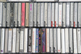 Rows of audio cassette tapes are tightly packed together. Many of the tapes have handwritten labels describing what is on the tape. 
