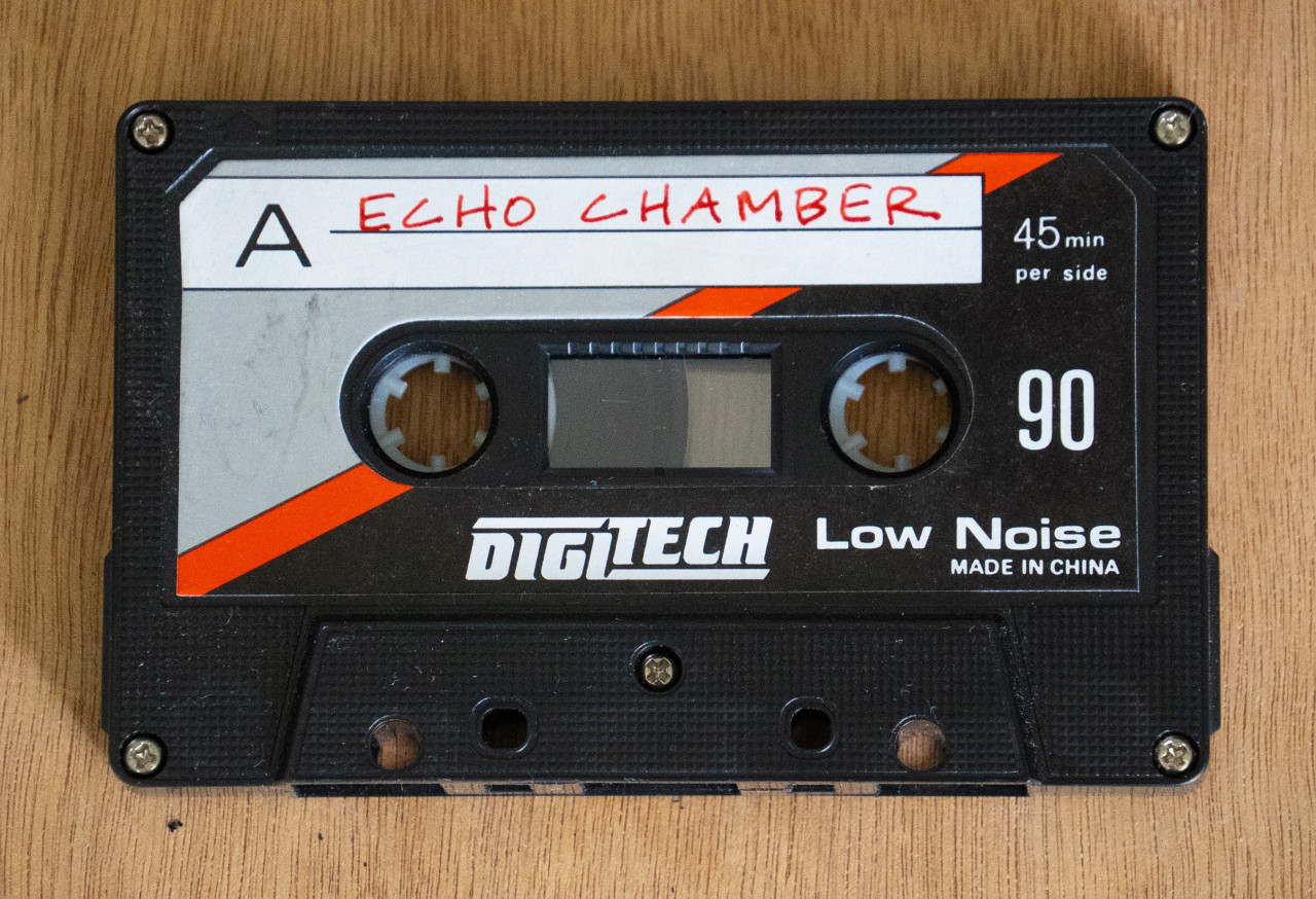 A black audio cassette tape with accents of grey and red on the cover. Handwritten text on the tape reads "ECHO CHAMBER." Behind the tape is a wooden background.