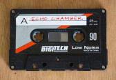 A black audio cassette tape with accents of grey and red on the cover. Handwritten text on the tape reads 