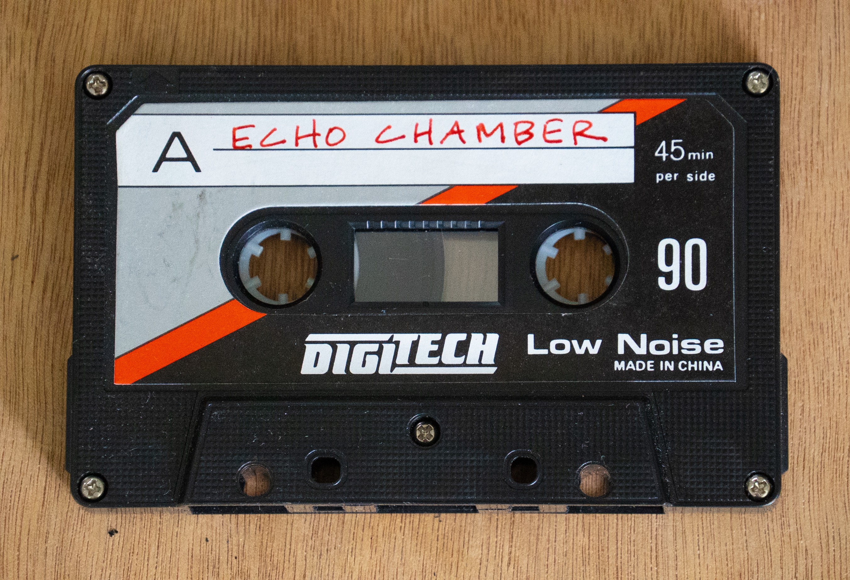 A black audio cassette tape with accents of grey and red on the cover. Handwritten text on the tape reads 