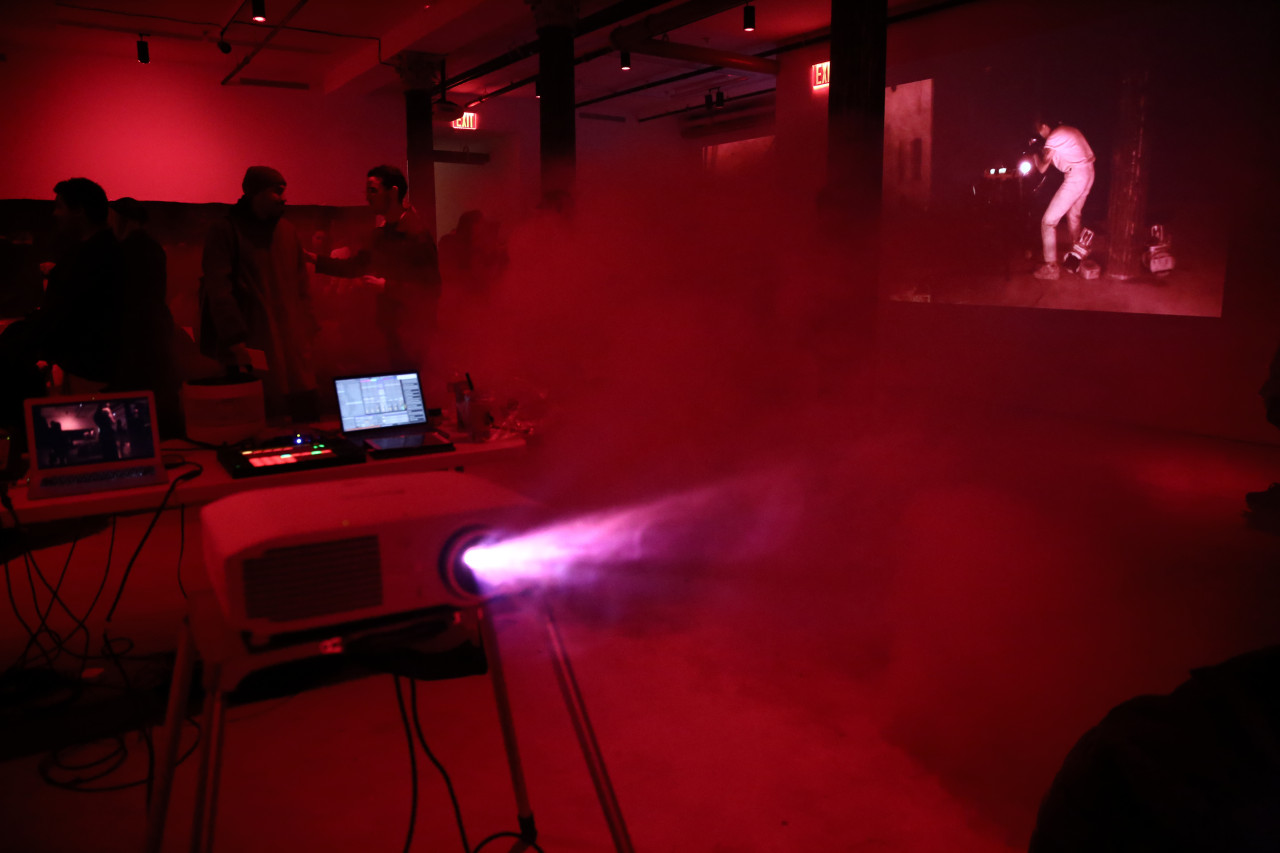 The bright beams of a projector cut through the fog filling a basement room. An image is projected onto one of the space’s walls. Several figures talk to each other in the background.
