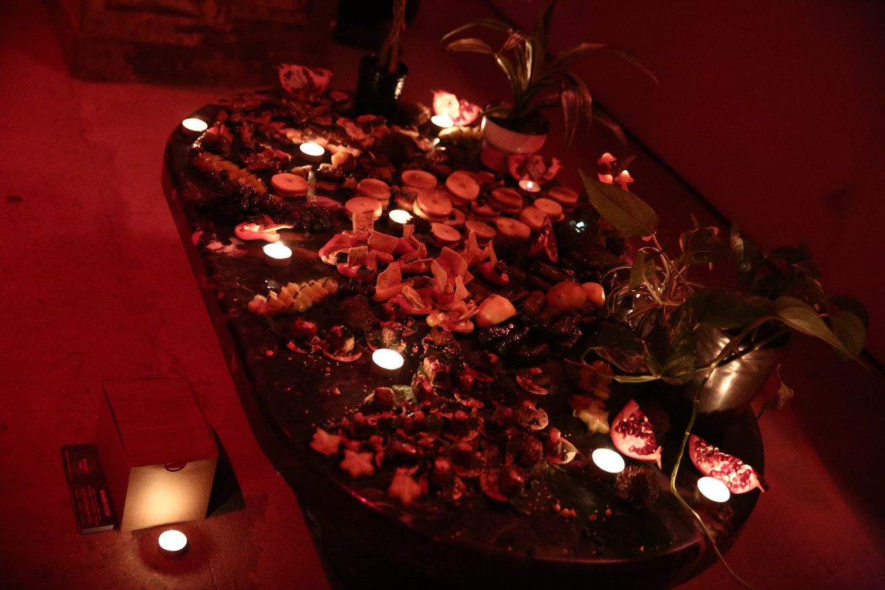 Half-eaten fruit, chocolate, candles, and grape leaves sit on a low, stone tabletop under dim lighting.