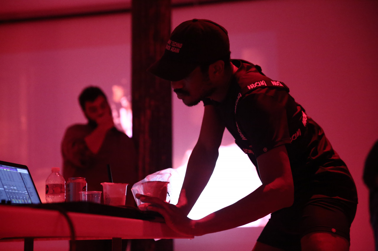 A figure in a baseball cap stands beside a table covered in scattered drinks and a laptop, typing on the computer’s keys.