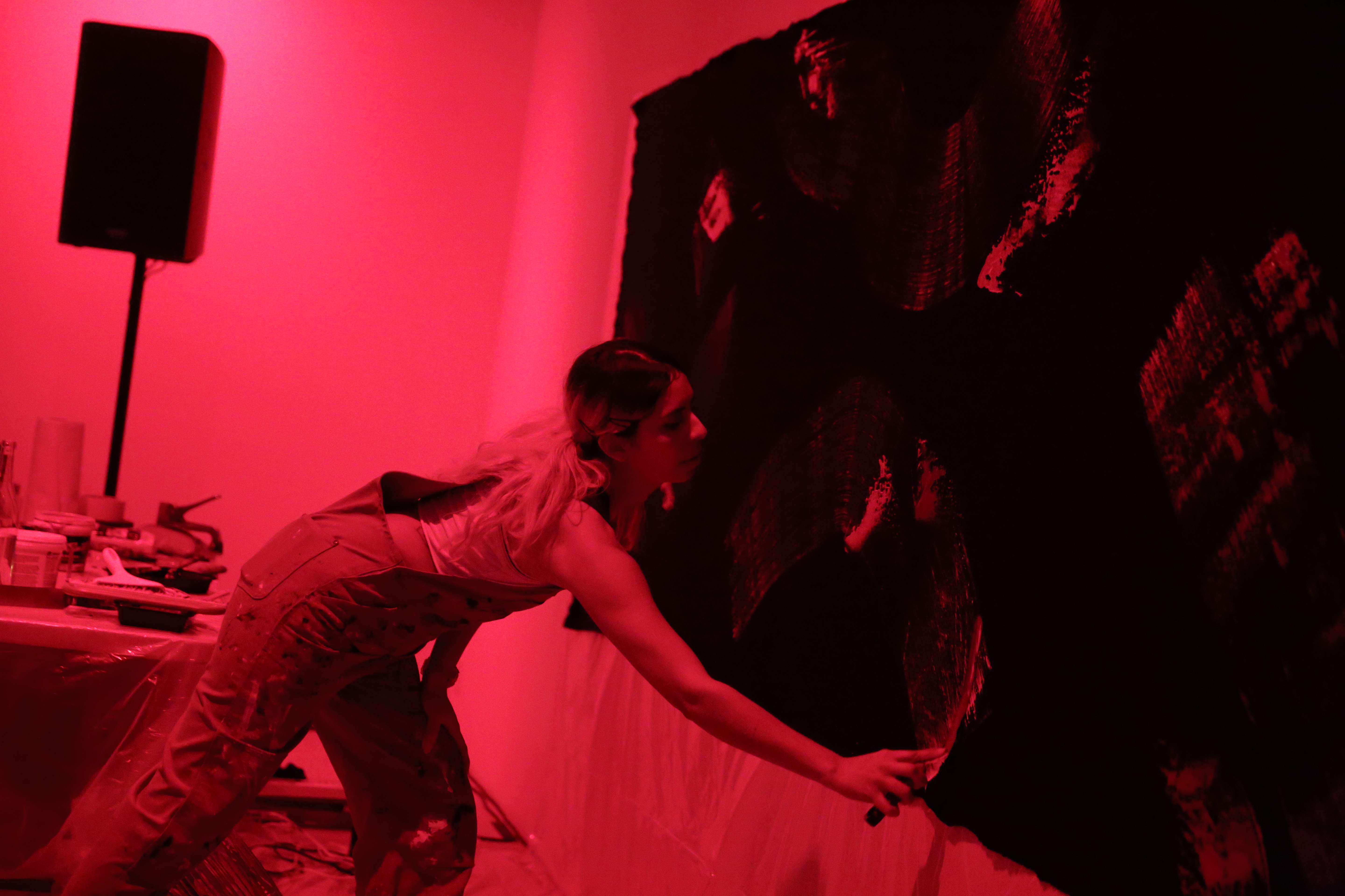 A figure in overalls leans forward to paint broad strokes of lighter paint on a black tarp hung on a wall. Painting supplies sit on a table directly behind them.