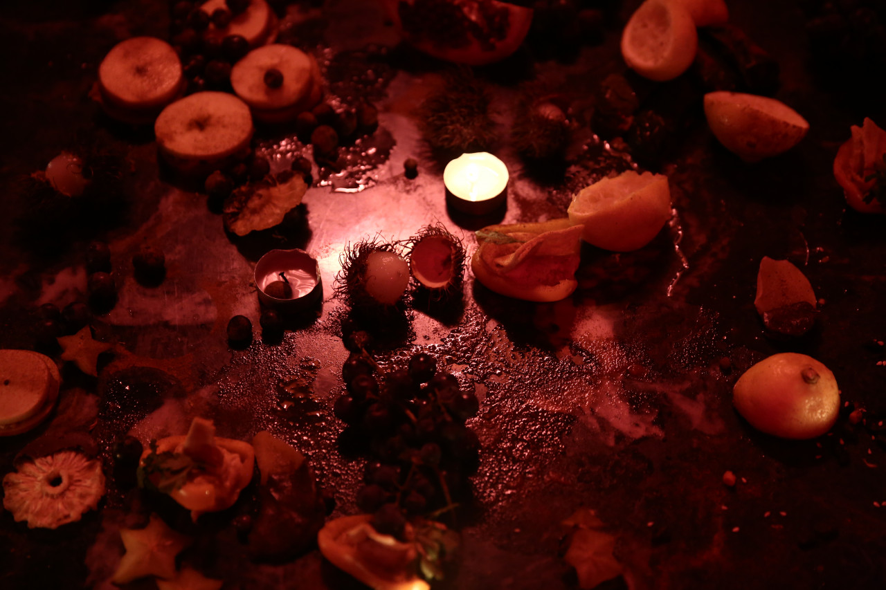 Half-eaten fruit, chocolate, and candles sit on a low, stone tabletop under dim lighting.