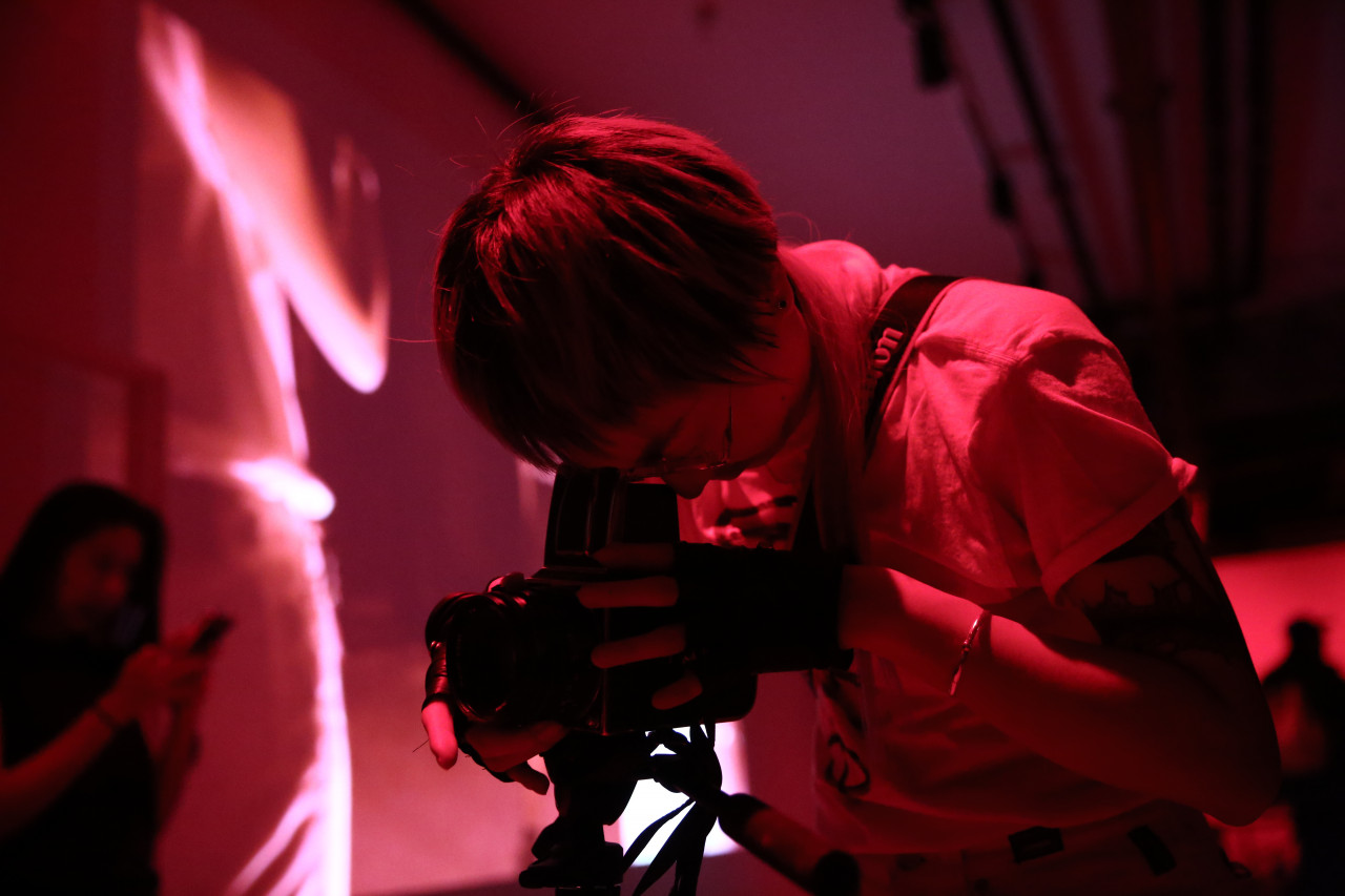 A figure looks into the viewfinder of a camera. In the background, an abstract image is projected onto a wall.