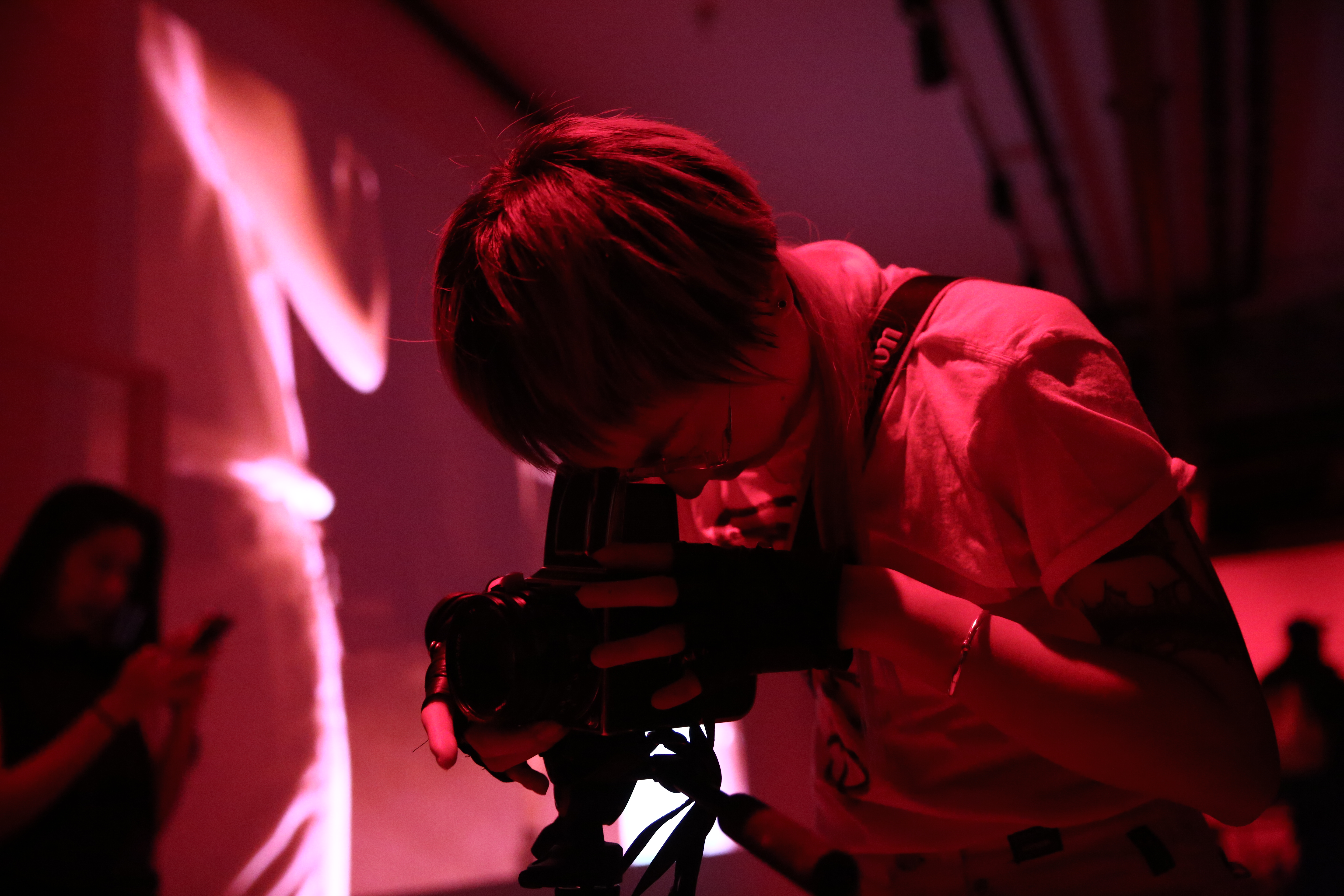 A figure looks into the viewfinder of a camera. In the background, an abstract image is projected onto a wall.