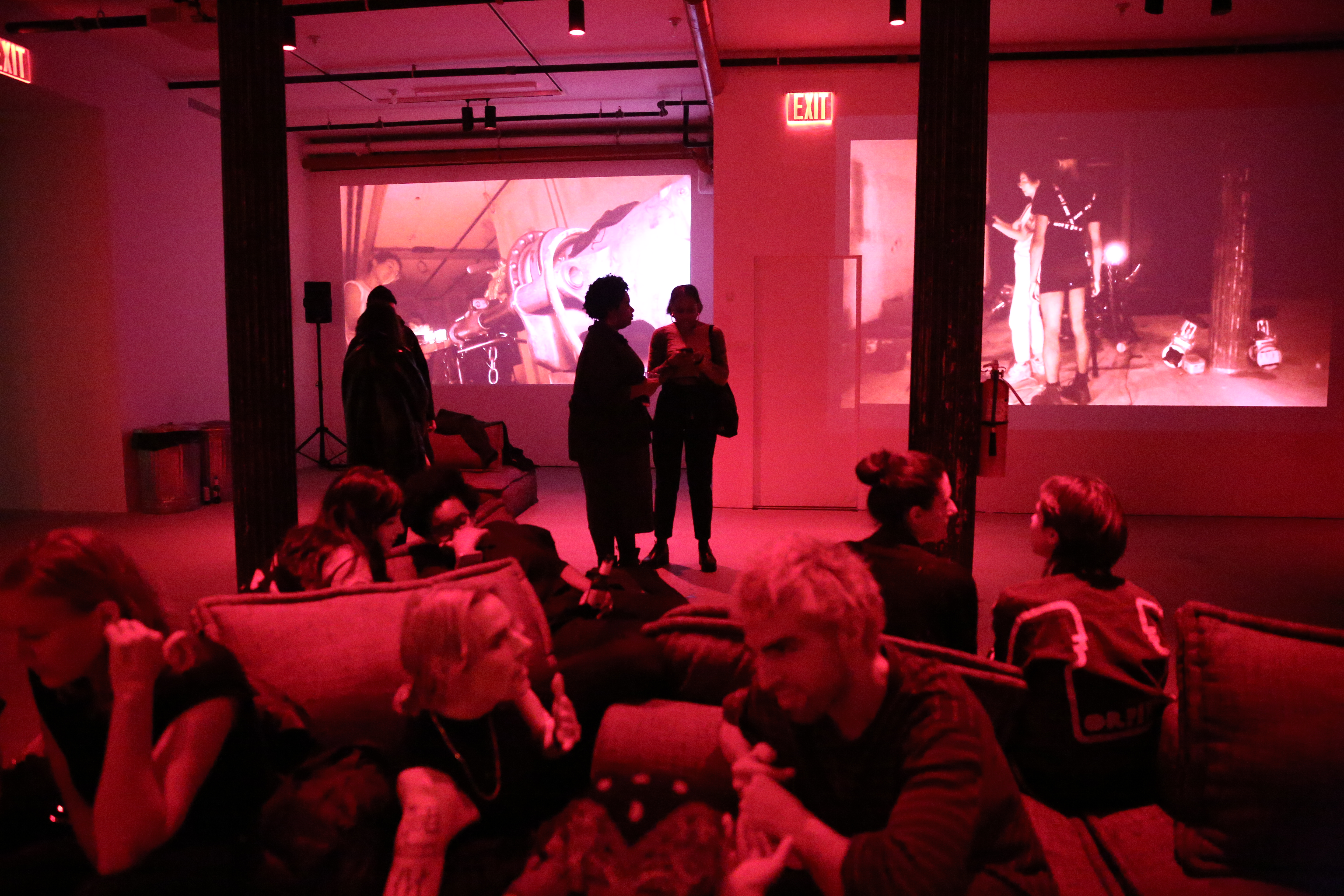 Several figures fill a dimly lit, basement room. Some figures sit and talk on large couches, while others stand and talk to each other. Video projections play on two of the space’s walls.