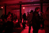 A crowd of people fill a dimly lit red room, some sitting and some standing. Abstract images are projected on the space’s walls.