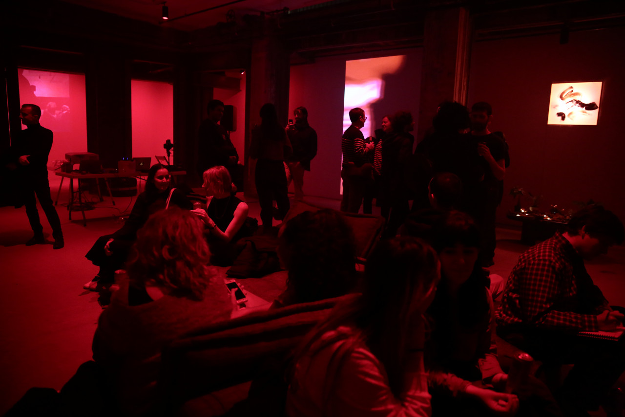 A crowd of people fill a dimly lit red room, some sitting and some standing. Projections of abstract images and an illuminated painting cover three of the space’s walls.