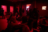 A crowd of people fill a dimly lit red room, some sitting and some standing. Projections of abstract images and an illuminated painting cover three of the space’s walls.