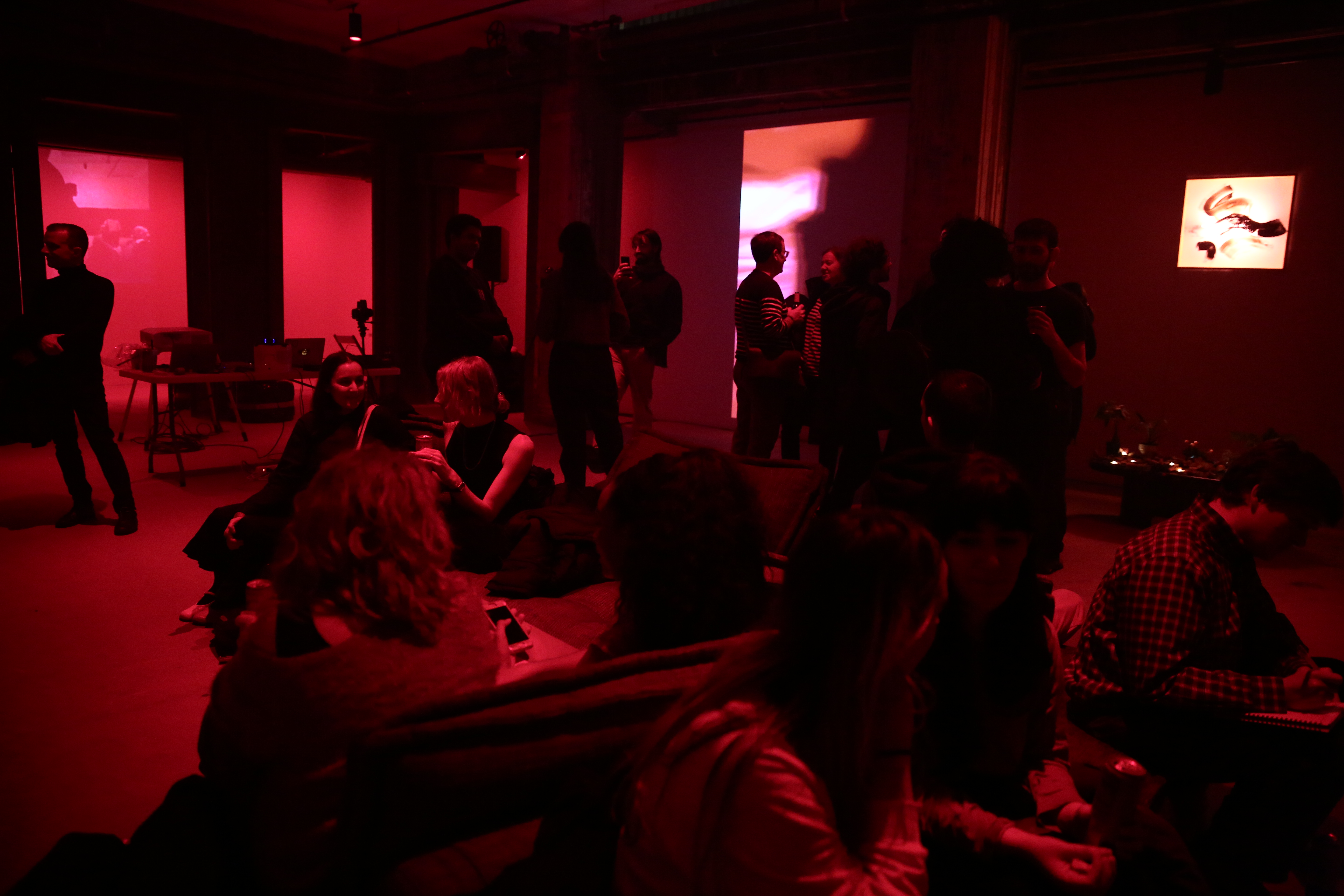 A crowd of people fill a dimly lit red room, some sitting and some standing. Projections of abstract images and an illuminated painting cover three of the space’s walls.