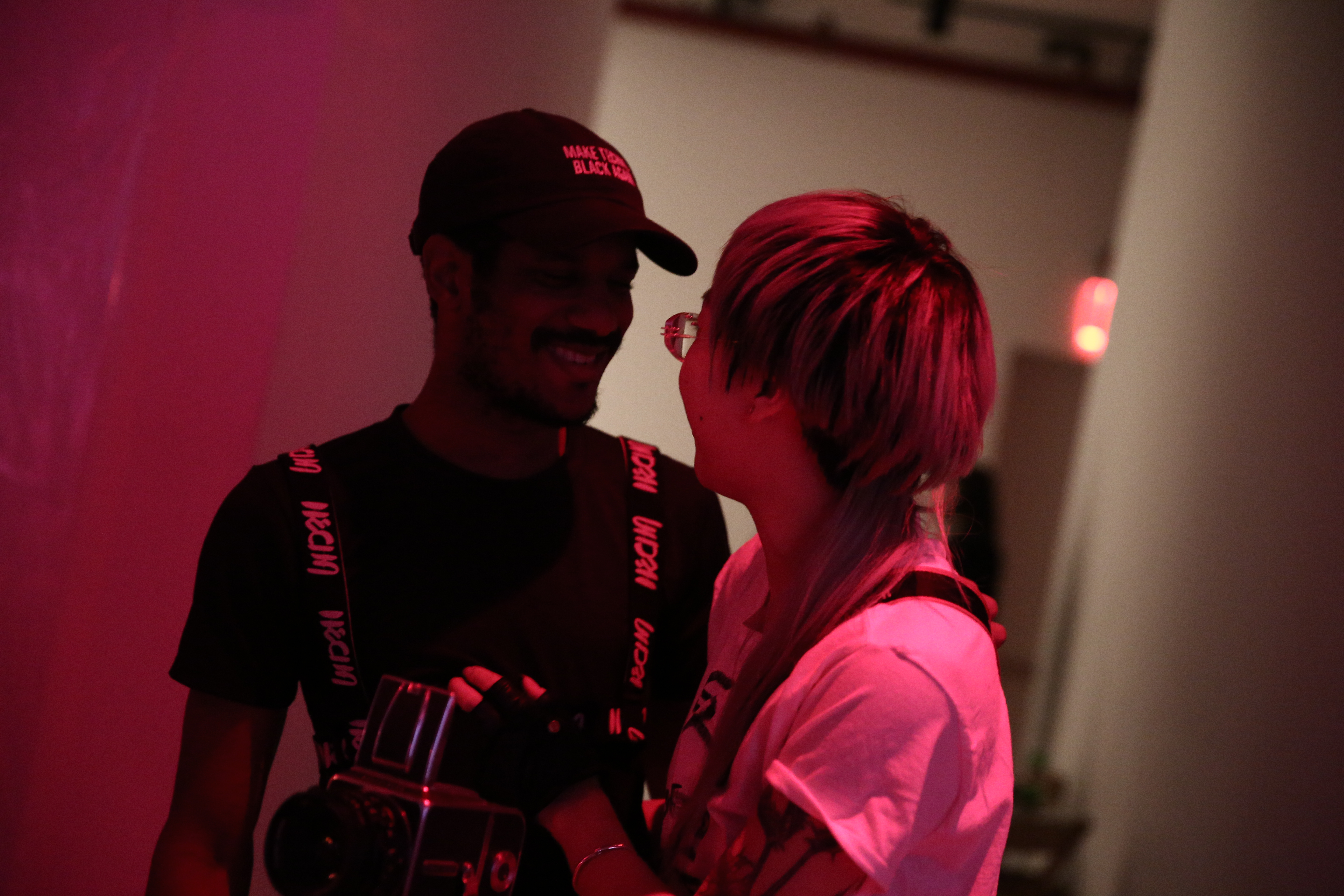 Two figures stand facing one another, smiling. One wears a baseball cap and the other holds a camera.