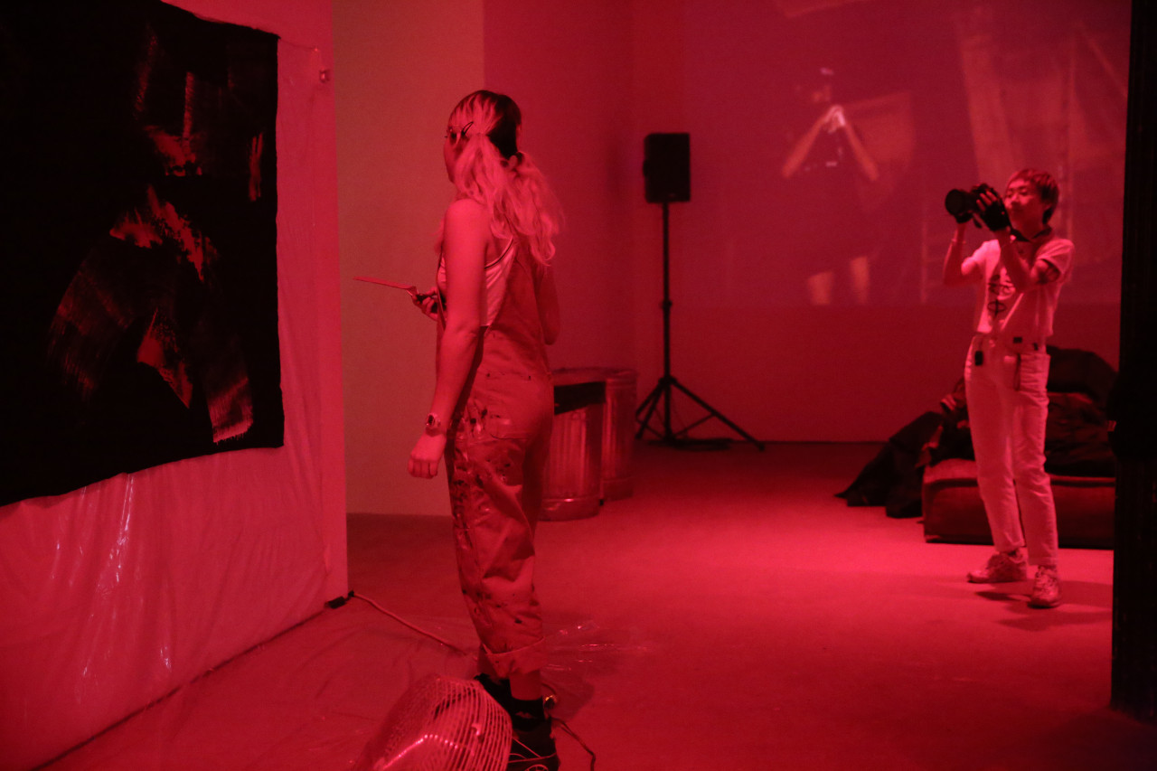 A figure stands facing a black tarp tacked to the wall. A second figure holds a camera, photographing the tarp. An image is projected on the back wall.