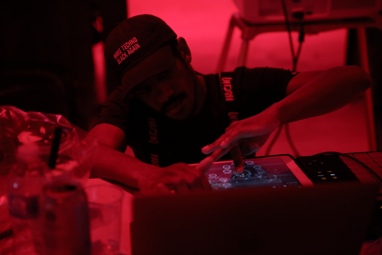 A figure kneels beside a table and traces their finger along the screen of an iPad. They wear a baseball cap that reads “MAKE TECHNO BLACK AGAIN.”