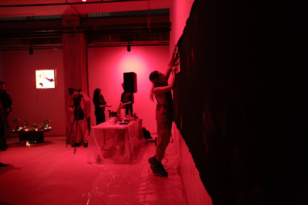 A figure in overalls reaches up to pin a dark tarp along the length of a wall. Behind them, another figure adjusts a camera on a tripod.