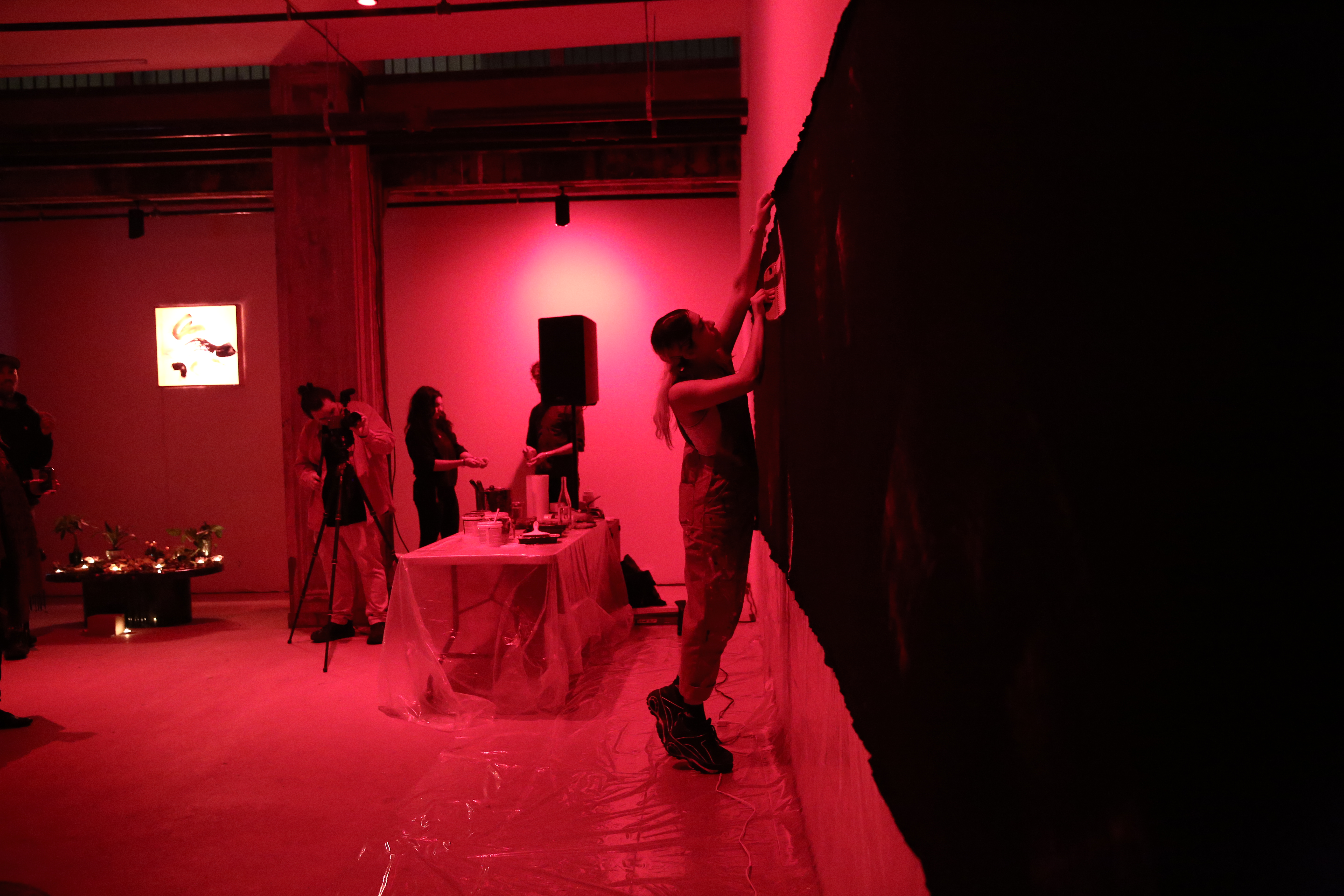 A figure in overalls reaches up to pin a dark tarp along the length of a wall. Behind them, another figure adjusts a camera on a tripod.