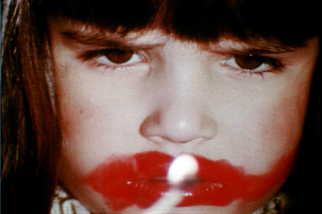 A closeup of a young girl with dark hair and bangs, frowning as she applies red lipstick in a messy circle around her mouth.