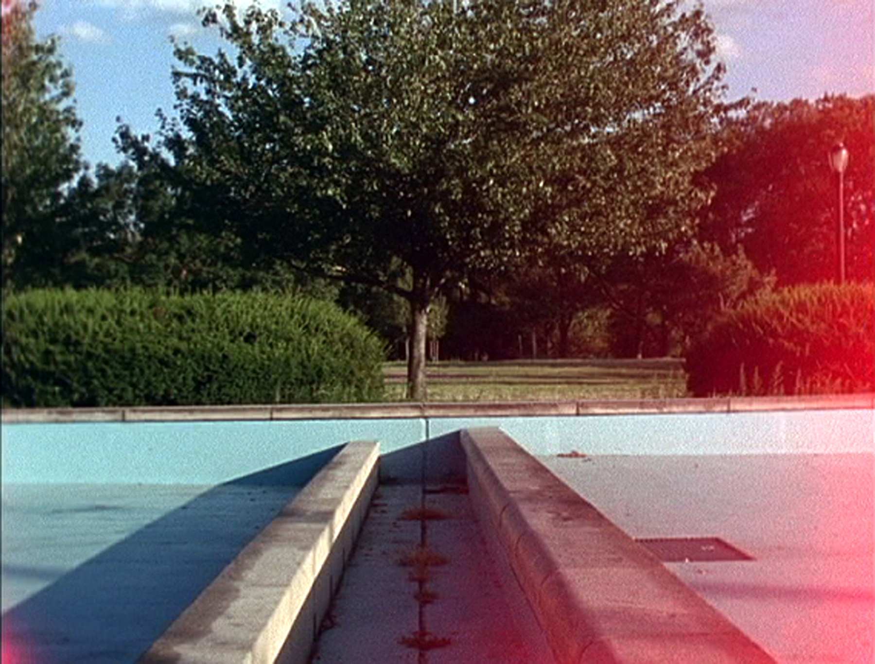 An empty pool with weeds growing out of cracks. There are trees and bushes in the background, and a pink glow on the righthand edge, as if from a sunspot.