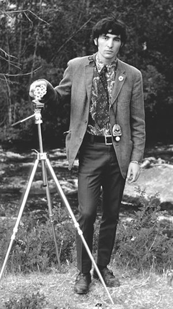 A black and white photograph of a man standing outside, surrounded by trees and grass, with his hand on a tripod.