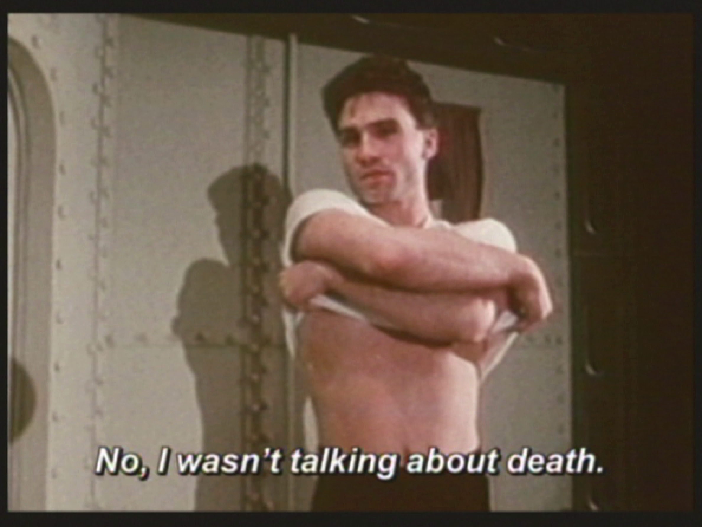 A man, looking toward the camera, in the midst of pulling his shirt off exposing his torso. A subtitle typed in white at the bottom reads 