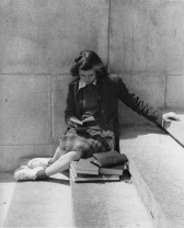A black and white image of a girl sitting sideways on stone steps against a wall reading a book. Several more books are piled in front of her.
