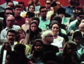A film still of a crowd of people sitting in rows of seats. Some wear sunglasses. Most do not speak to one another but gaze forward or glance around themselves.