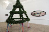 A framed, oval-shaped painting of a man and a conical plant sculpture decorated with hanging crepe paper tassels are displayed in a gallery.