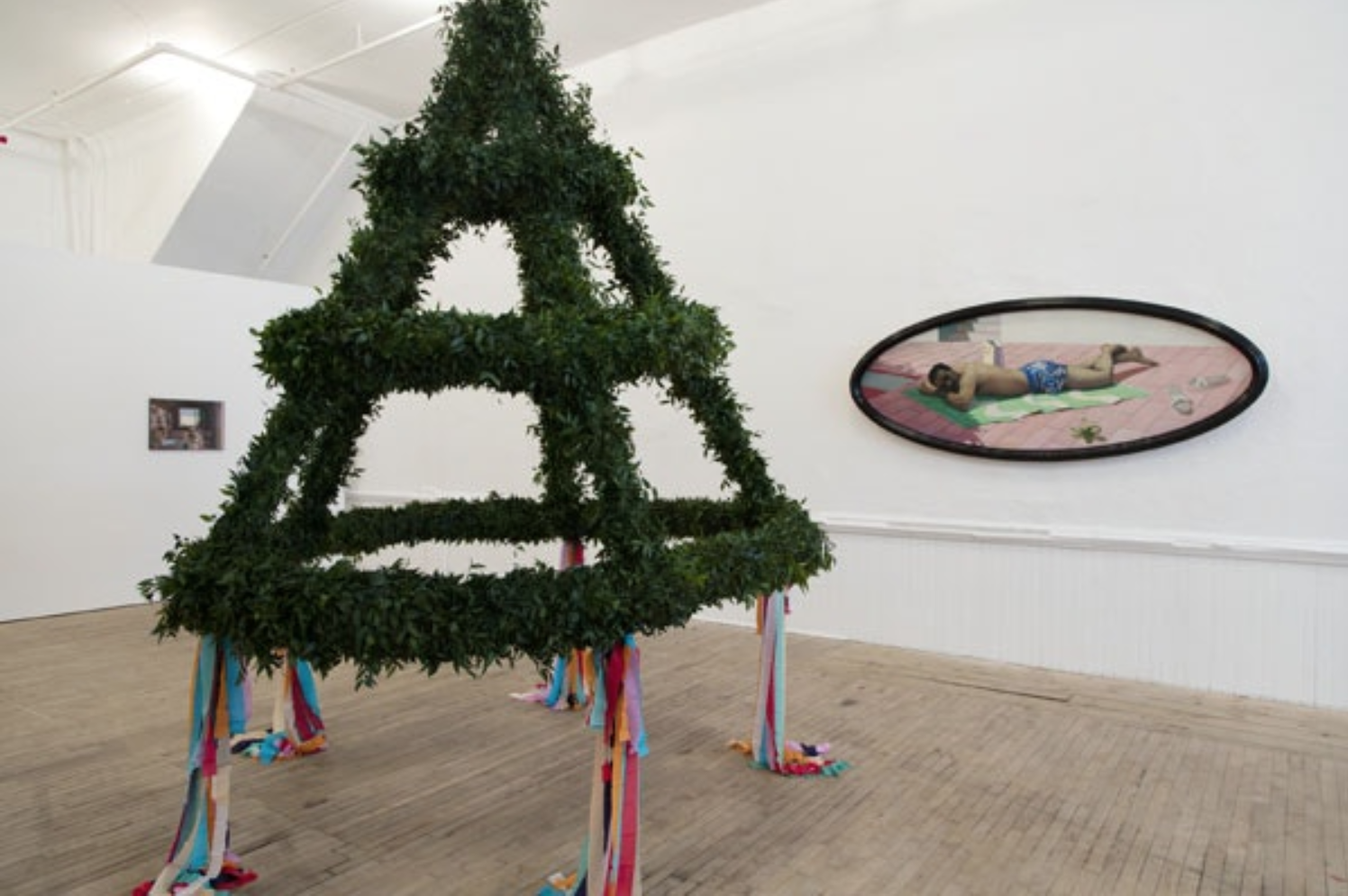 A framed, oval-shaped painting of a man and a conical plant sculpture decorated with hanging crepe paper tassels are displayed in a gallery.