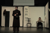 A person is standing at the end of a stage, dressed in a long coat, with a stern expression on her face. A man sits at a desk in the corner, his head in his hands. There is a white trifold screen behind them, with an extra chair, and a banner written in German.