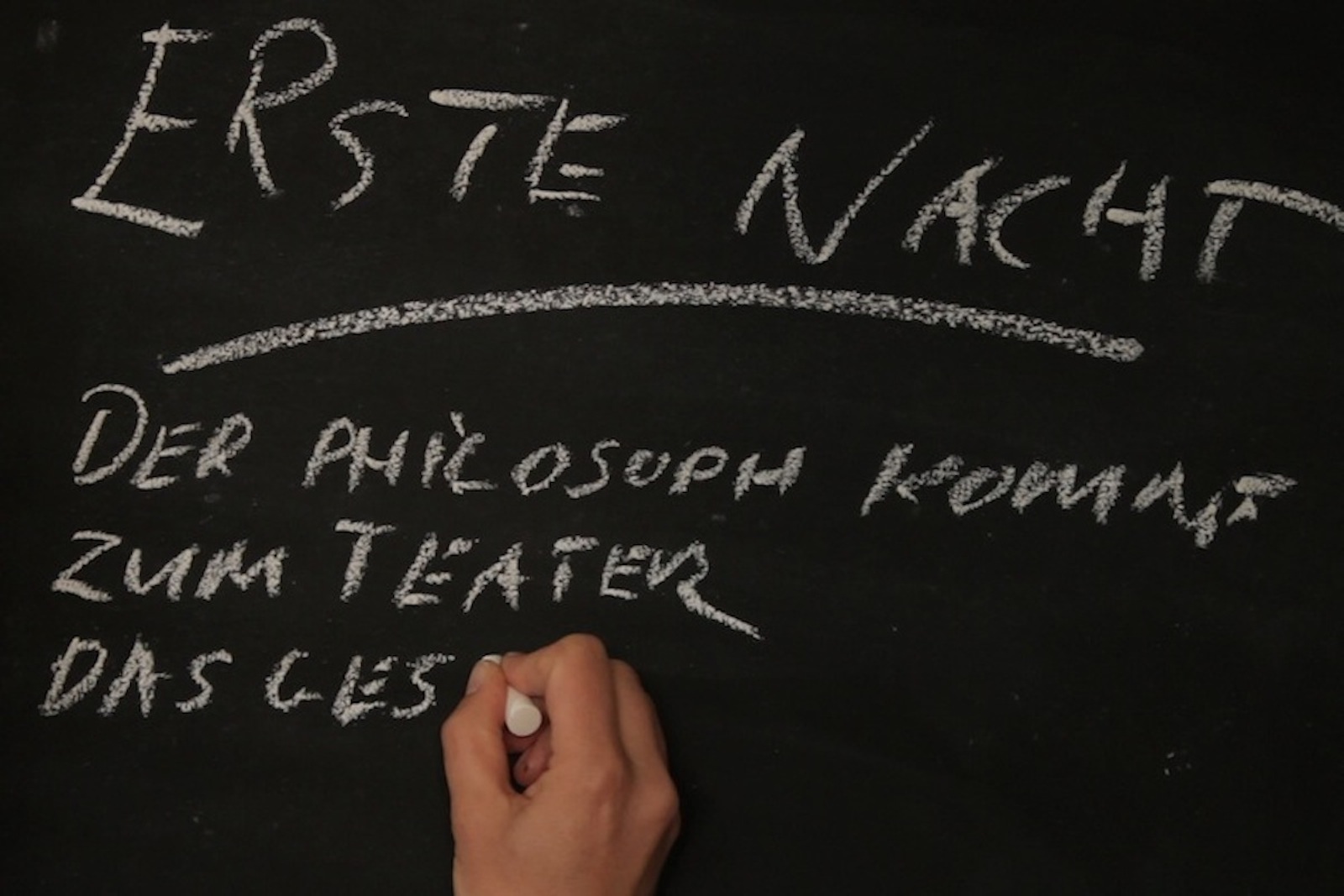 A hand is writing in German on a chalkboard with white chalk.