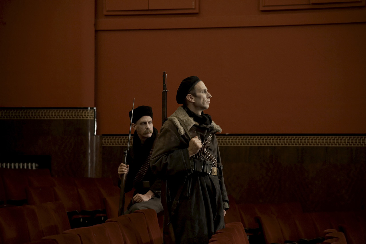 Two men dressed in uniform and carrying guns are looking toward the right of the image. They are within the red seats of a theatre; one is sitting and one is standing.