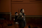 Two men dressed in uniform and carrying guns are looking toward the right of the image. They are within the red seats of a theatre; one is sitting and one is standing.
