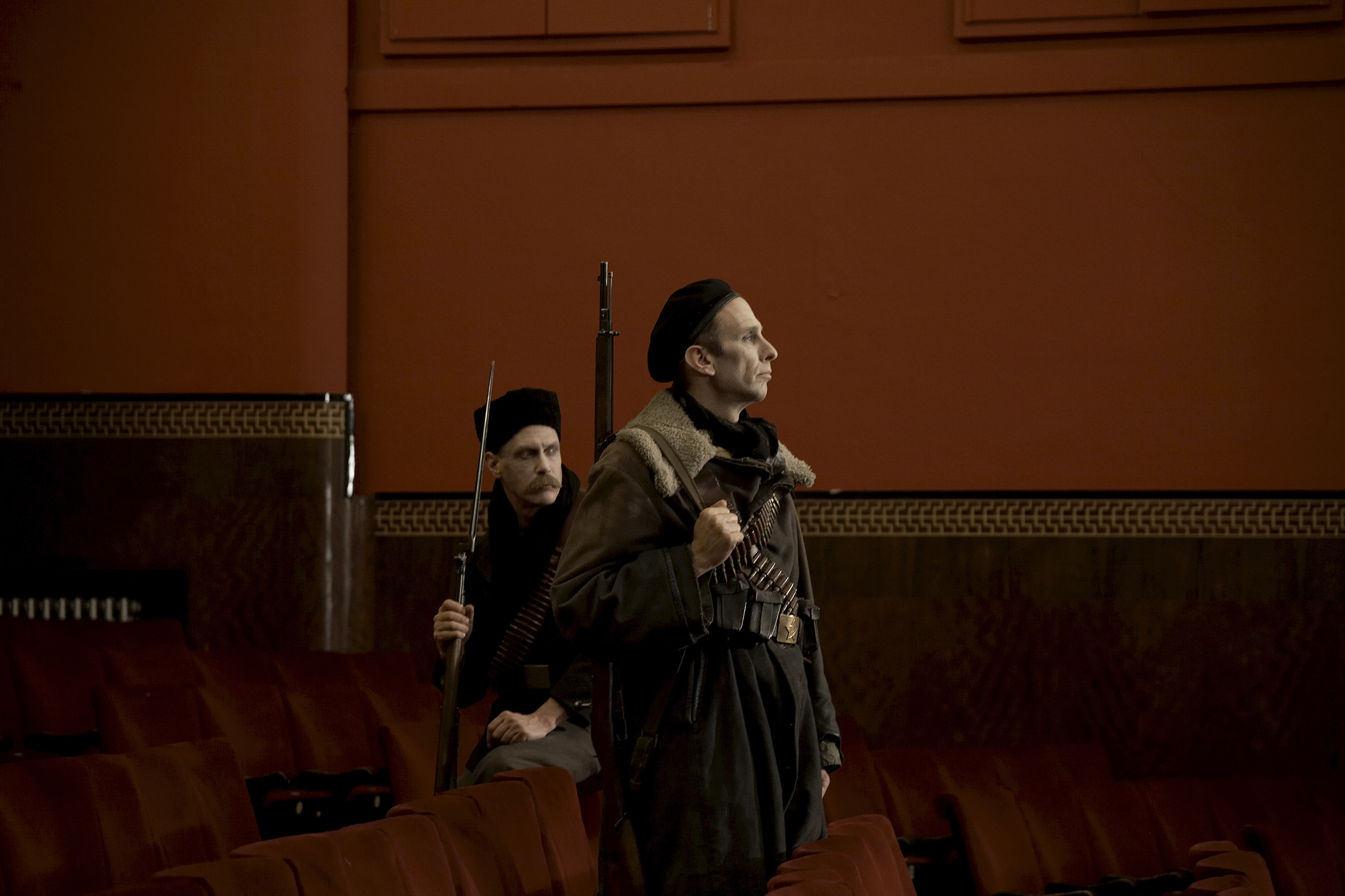 Two men dressed in uniform and carrying guns are looking toward the right of the image. They are within the red seats of a theatre; one is sitting and one is standing.