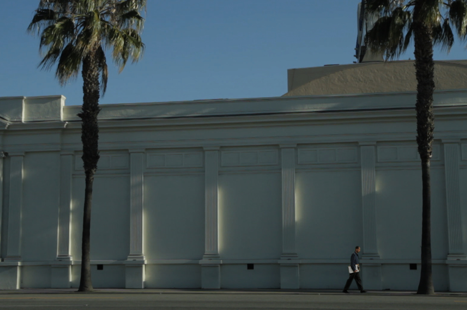 A man holding a clipboard at his side walks past a large, columned white wall with palm trees on either end.