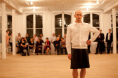 A man with a shaved head stands in front of a crowd seated at a long table in a large gallery space. He stares in front of him with one hand resting at his hip and wears a white collared shirt and a black, knee-length skirt.