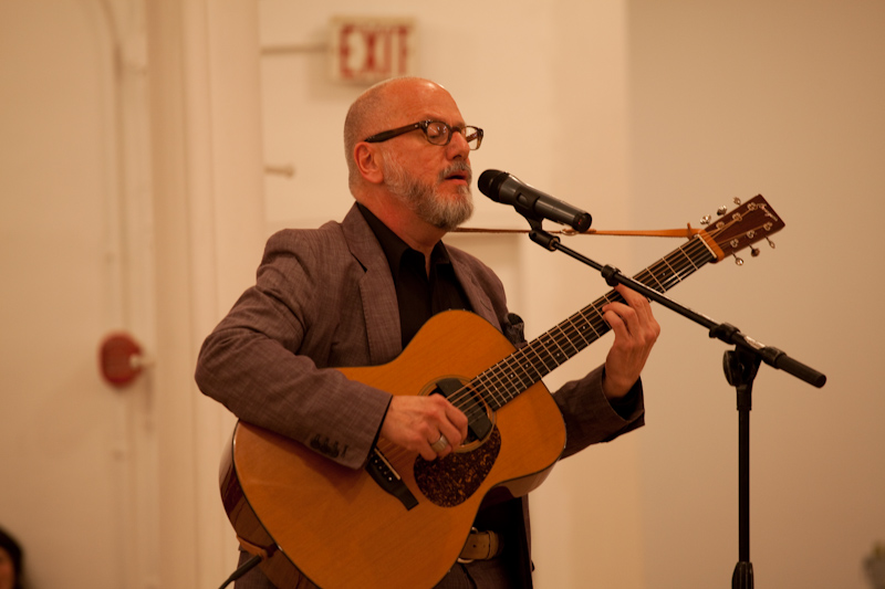 A man in a suit, black shirt, and glasses strums a guitar while singing into a microphone in a white-walled gallery space.