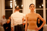 A woman in a nude leotard-like top and silver leggings stands facing forward with her hand on her hip in a gallery space. Behind her, a figure in a black skirt and white collared shirt stands in a similar position, with his back to the former performer.