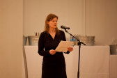 A woman in a dark dress with shoulder-length auburn hair stands before a microphone in a gallery space, reading from a sheet of paper. She looks up from the paper to gaze in front of her.