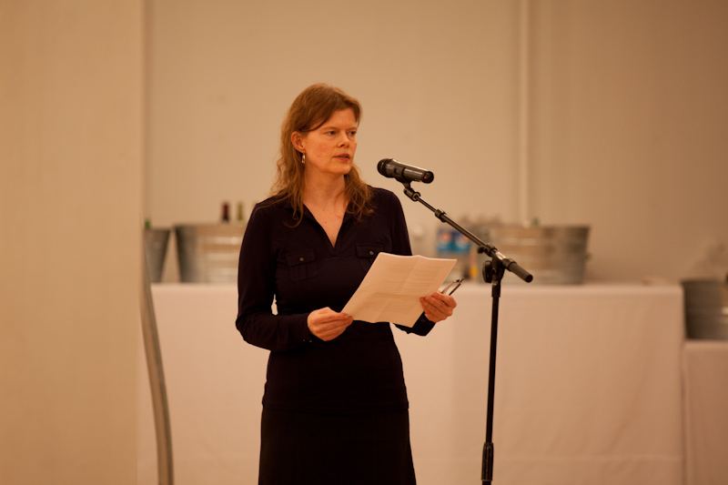 A woman in a dark dress with shoulder-length auburn hair stands before a microphone in a gallery space, reading from a sheet of paper. She looks up from the paper to gaze in front of her.