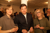 Three figures pose for a photograph in a gallery space crowded with milling people who talk amongst each other. They hold drink glasses in their hands and are illuminated by the camera's flash.